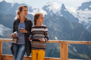 Mom with daughter drinking warm tea in the rustick wooden terrace on mountain, alpine view, snow on hills. Winter weekend. Dombay, Karachay-Cherkessia, Caucasus, Russia.